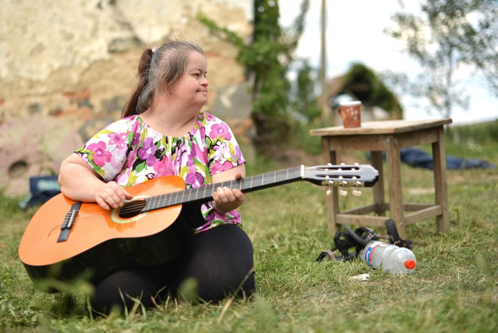 Woman with floral pattern shirt sitting on the grass with a guitar.